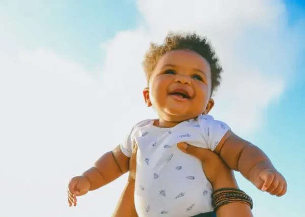 African American baby in spotty top and blue jeans held aloft to the clouds at the beach.